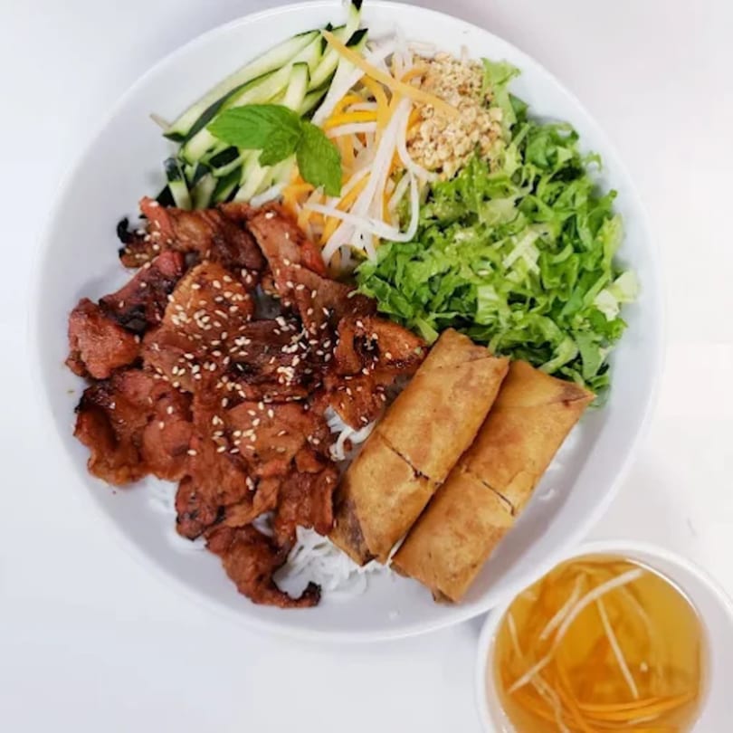 Vietnamese vermicelli bowl with grilled pork, spring rolls, fresh vegetables, and a side of sauce.