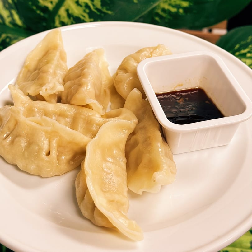 A plate of steamed dumplings with a side of soy sauce, against a green leafy background.