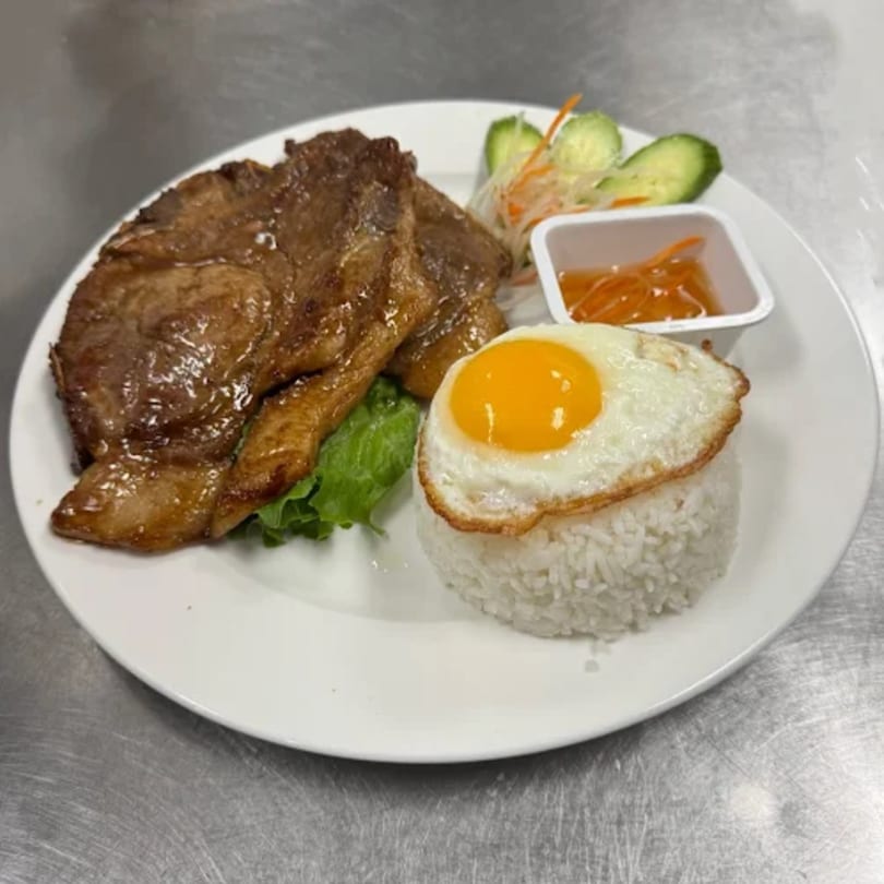 Vietnamese grilled pork chop with a fried egg, rice, and pickled vegetables on a white plate.